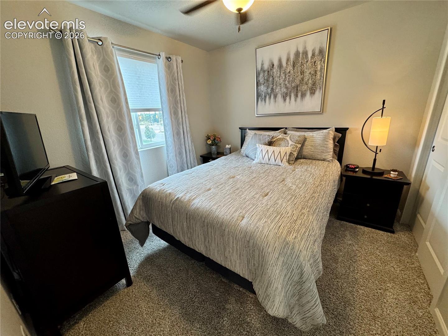 Neutral-toned bedroom featuring carpeting, a ceiling fan with light fixture, and a single window with rod-mounted drapery