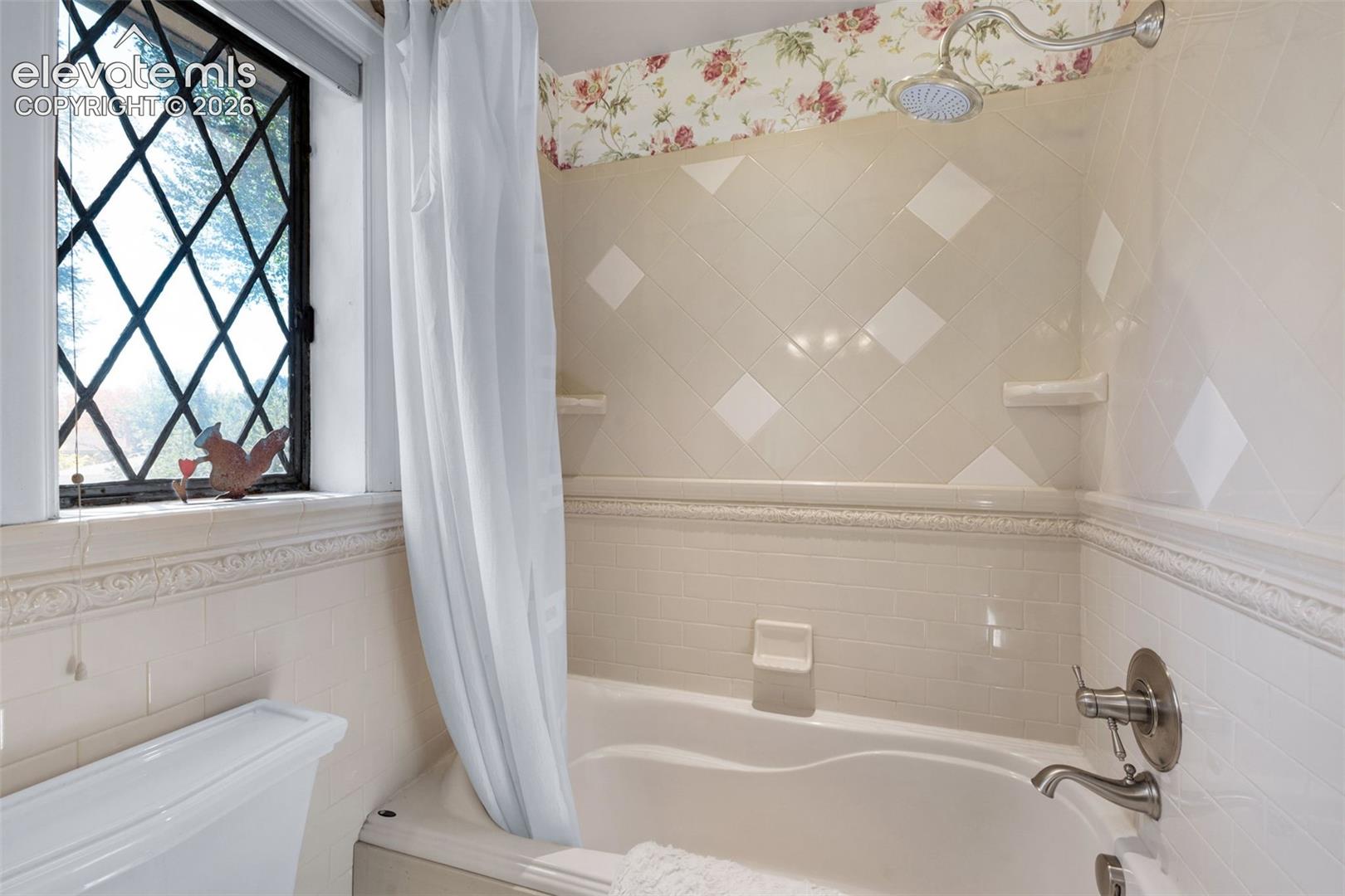 Bathtub enclosure featuring diamond-patterned tile, integrated tile soap niches, decorative crown molding trim, and a leaded glass window