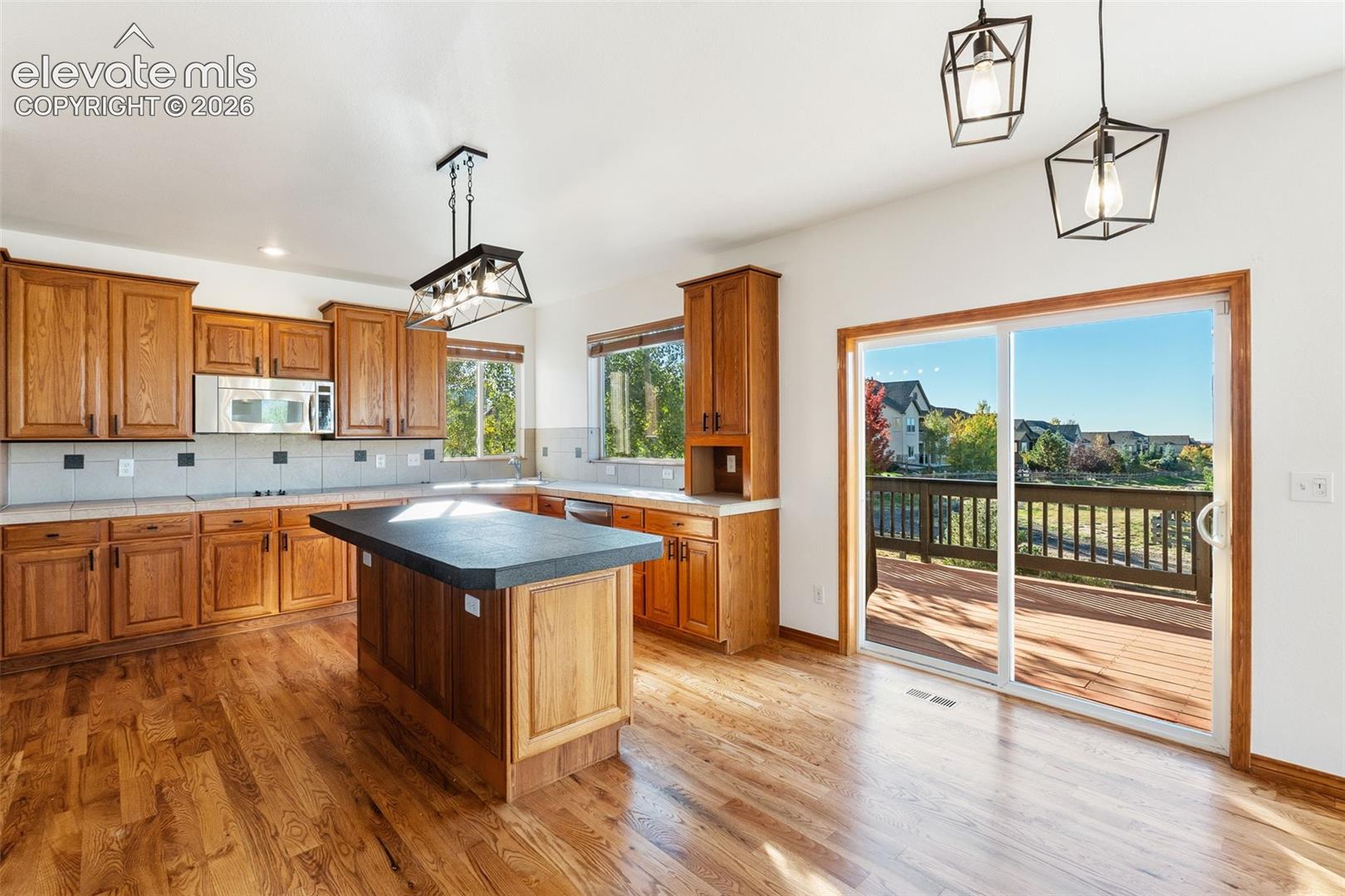 Kitchen featuring wood cabinetry, central island with a dark solid surface countertop, wood-finish flooring, and metal geometric pendant lighting, with sliding glass doors opening to a wood deck