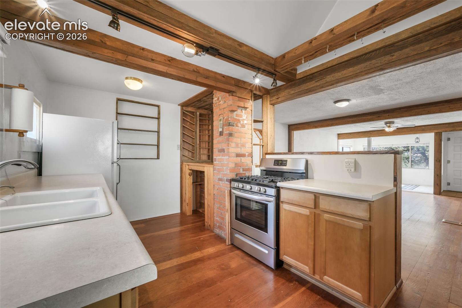 Kitchen featuring wood-finish flooring, exposed wood ceiling beams, stainless steel gas range, light-toned cabinetry, and a brick feature wall