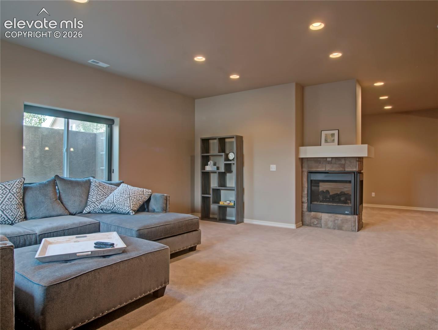 Spacious above grade living area featuring bright light, a two sided fireplace with a stone surround and mantel, recessed lighting, neutral carpet, and many windows providing natural light. 