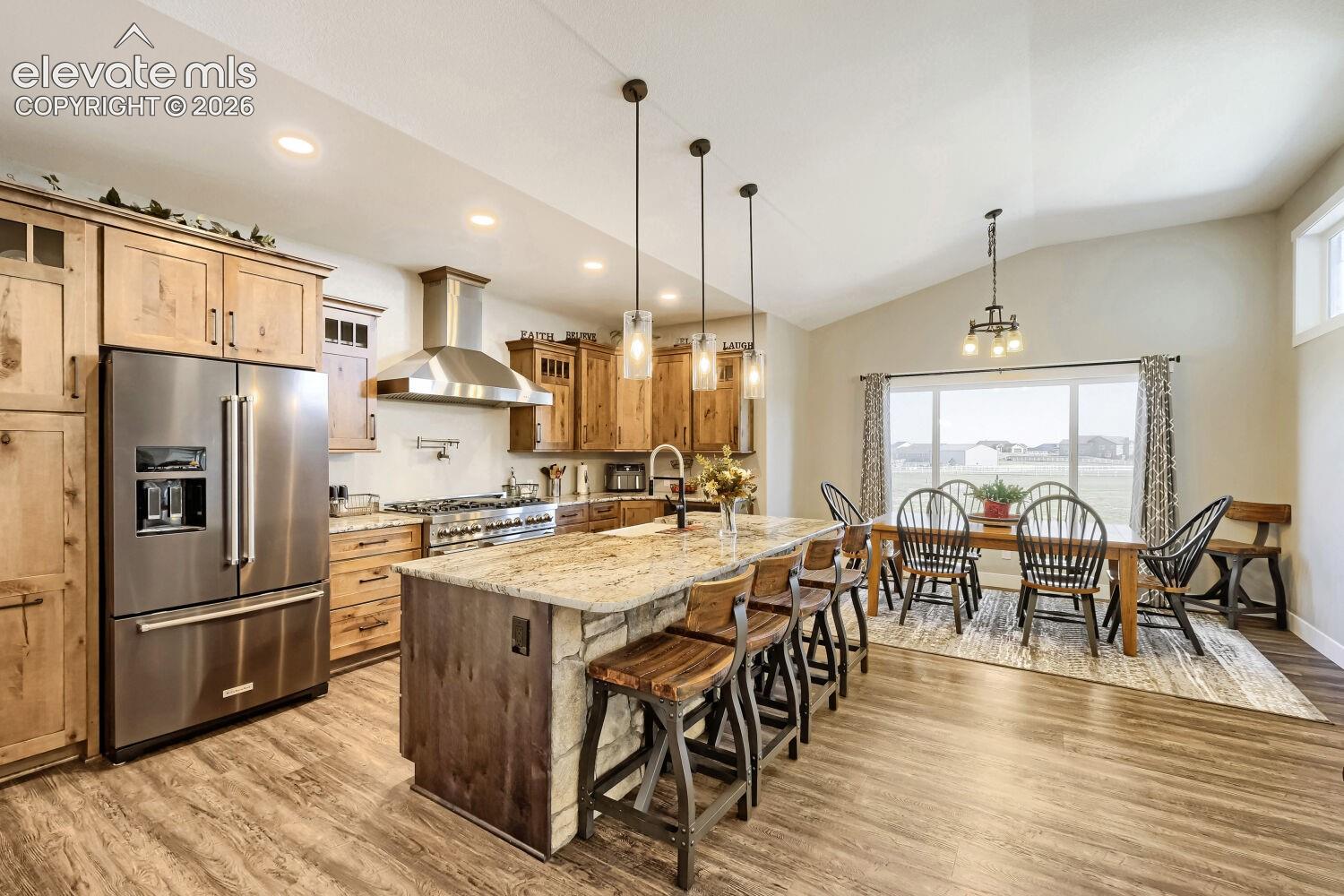 Vaulted ceiling, wood-finish flooring, stone-accented kitchen island with pendant lighting, stainless steel appliances, and wood cabinetry