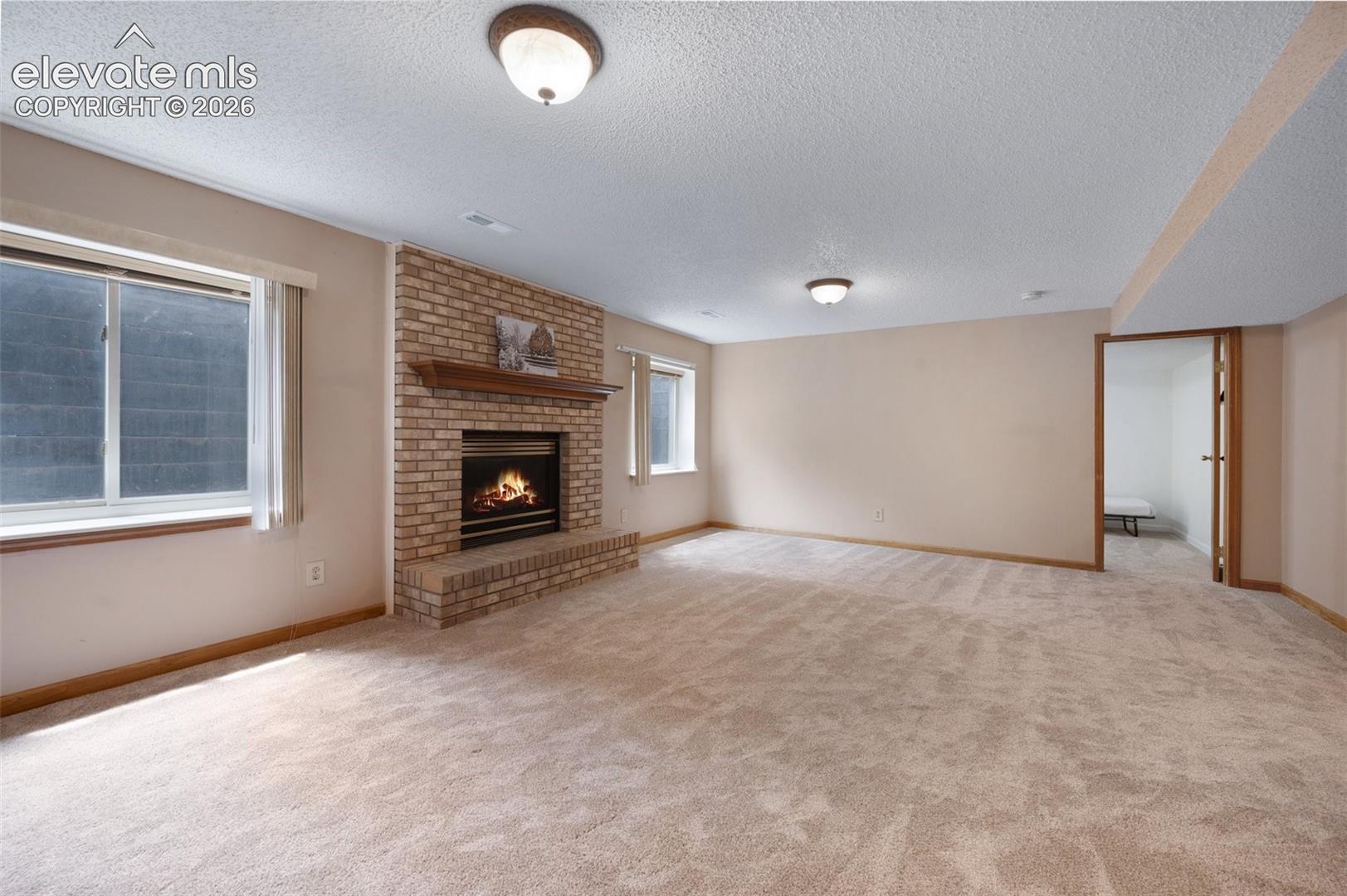 Unfurnished living room featuring a textured ceiling, carpet flooring, a brick fireplace, and baseboards. Fire in fireplace staged and added.