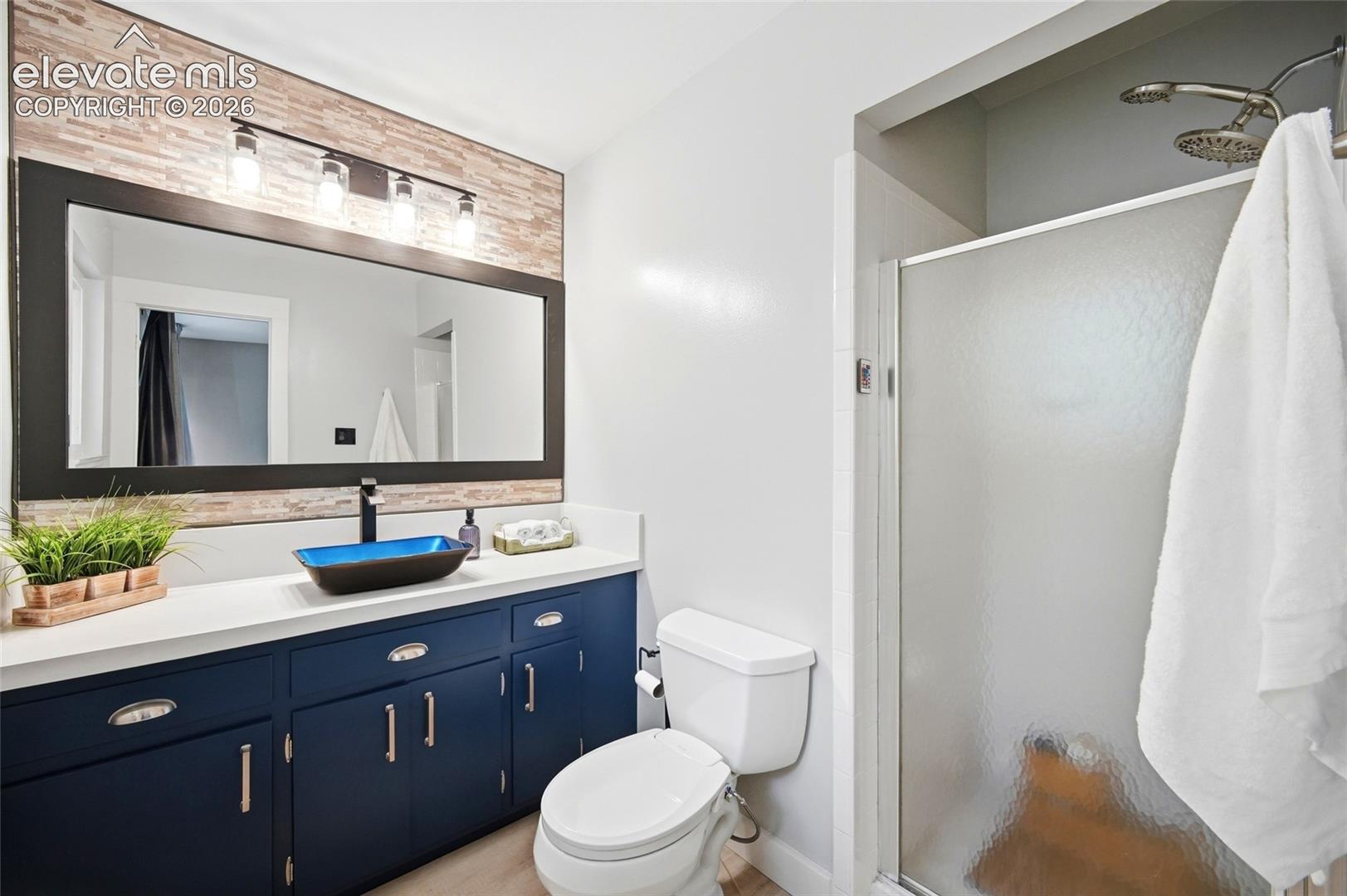 Navy blue vanity cabinet with solid surface countertop, rectangular vessel sink, framed mirror with integrated light fixture, and walk-in shower with semi-opaque glass door