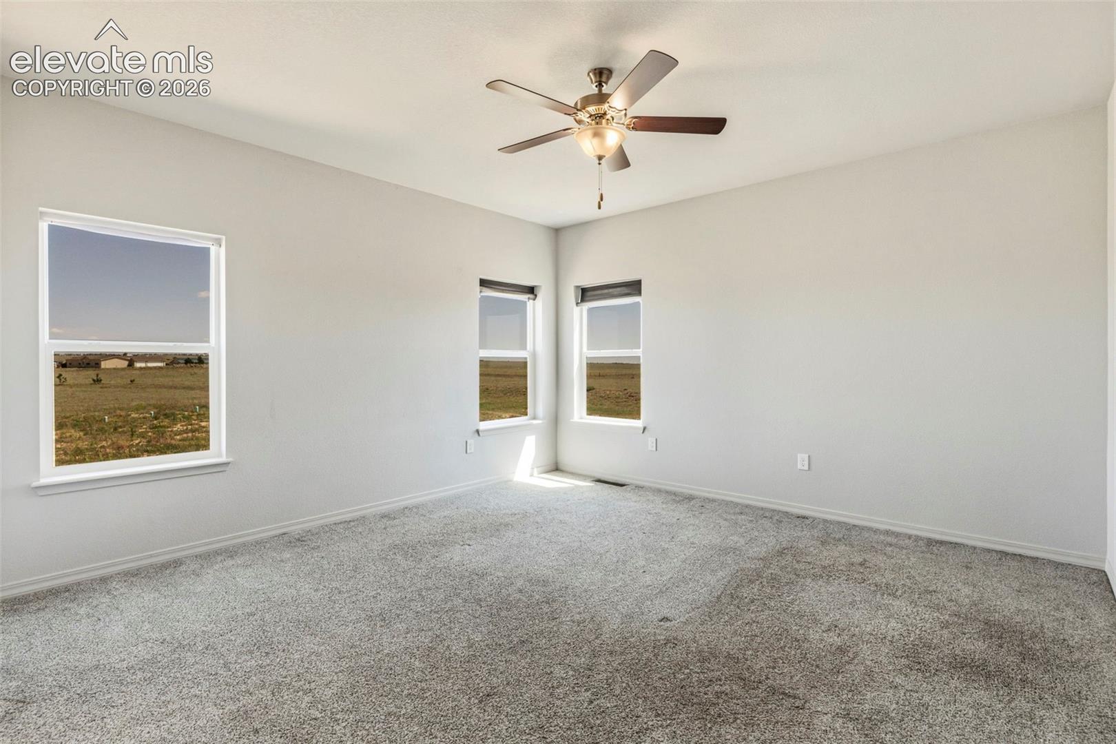 Spacious room featuring light gray carpeting, multiple windows providing natural light, a ceiling fan with integrated lighting, and neutral wall tones