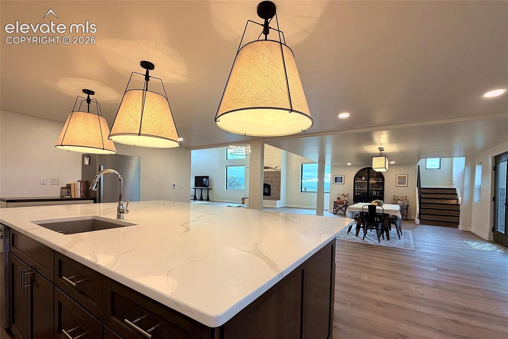 Kitchen island featuring a white surface with grey veining, dark cabinetry, recessed lighting, and trio of pendant light fixtures with fabric shades, complemented by wood-finish flooring