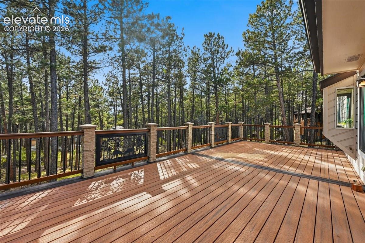 Expansive wood-finish deck with decorative metal railing panels and stone-wrapped posts, surrounded by mature trees