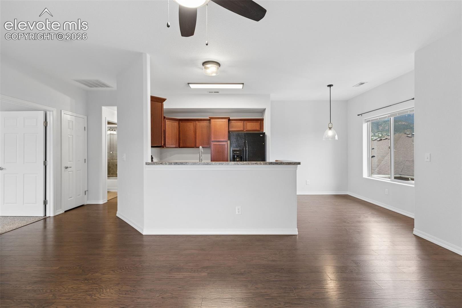 Open-concept interior featuring wood-finish flooring, a kitchen peninsula with a stone-surface countertop, cabinetry with a rich wood finish, and a ceiling fan with integrated lighting