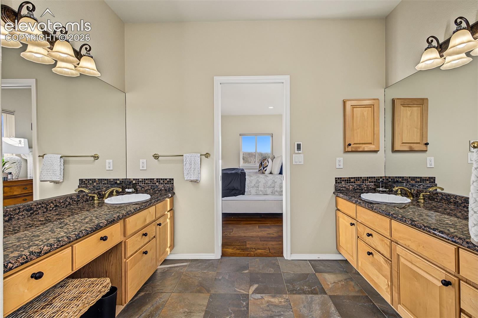 Spacious primary bathroom featuring separate vanities with granite countertops, wood cabinetry, decorative lighting, and tile flooring.