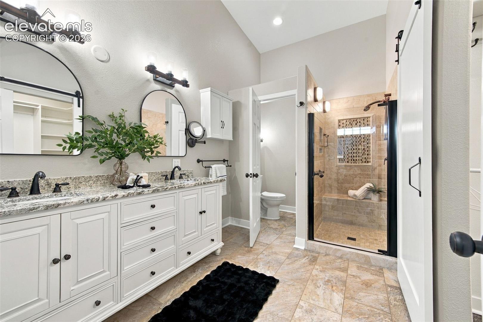 Dual-vanity cabinetry with stone countertops, arched wall mirrors, vaulted ceilings, and a walk-in shower with stone tile surround and built-in bench
