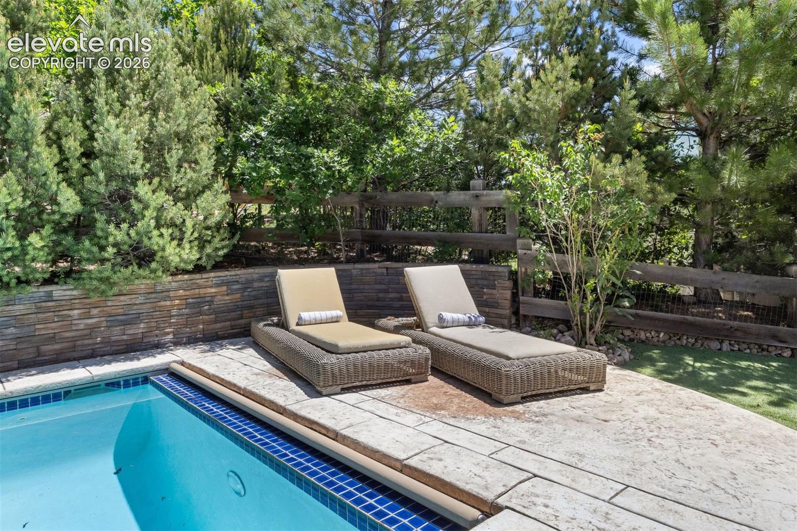 Swimming pool with blue tile coping, stamped concrete decking, a stacked stone retaining wall, and mature landscaping
