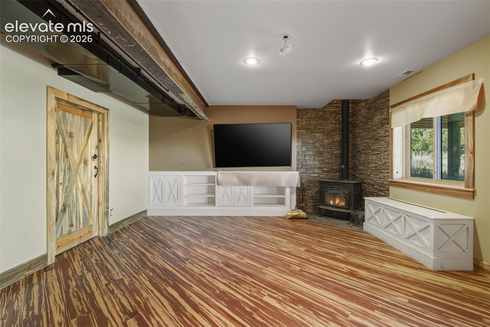 Wood-finish flooring, built-in white cabinetry, stone veneer accent wall, wood-burning stove, and a window with wood trim