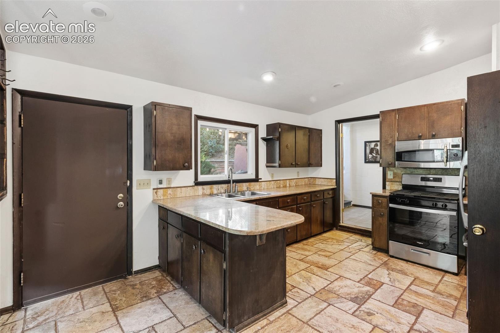 Kitchen featuring dark wood cabinetry, stone-style tile flooring, stone countertops, and stainless steel appliances