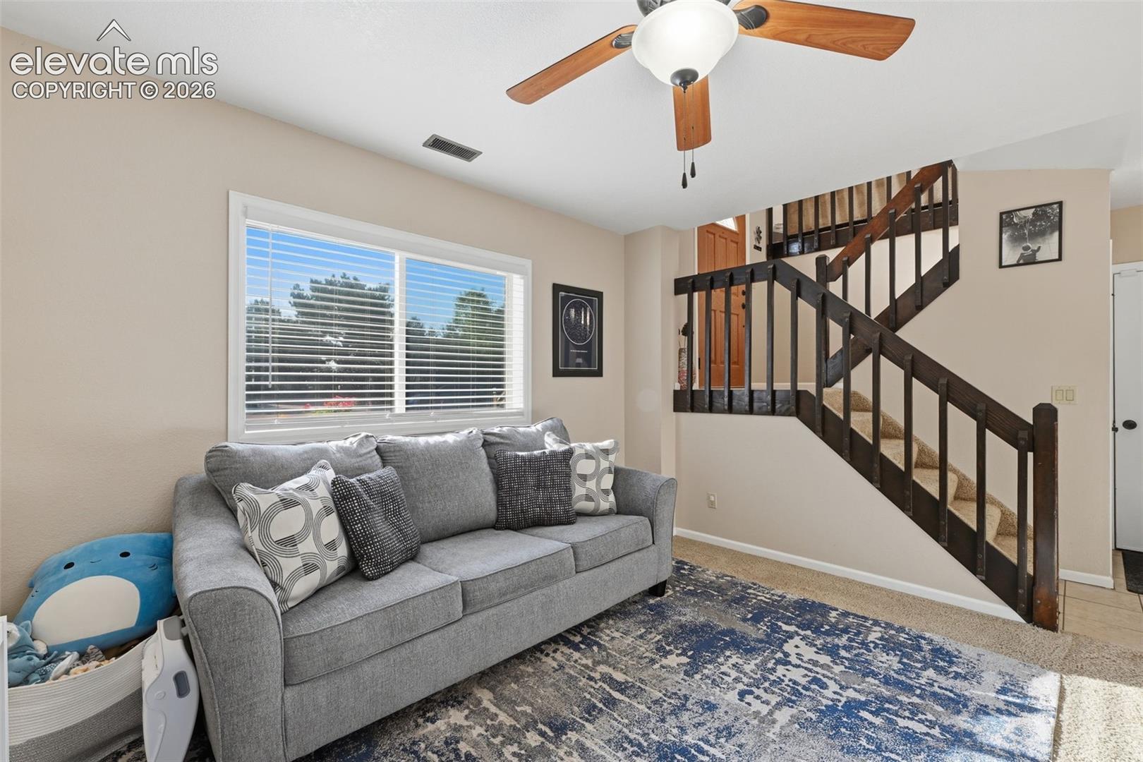 Living area featuring a large window with blinds, a ceiling fan with light fixture, carpeted stairs with wood railings, and neutral wall tones