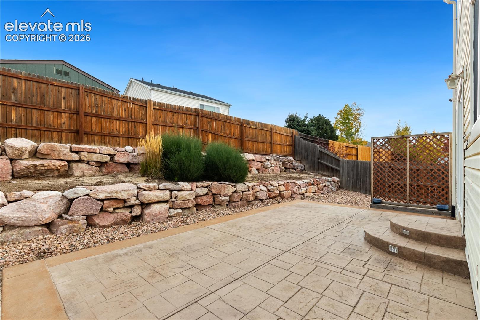 Stamped concrete patio featuring tiered natural stone retaining walls, landscape plantings, wood-style privacy fencing, and recessed step lighting