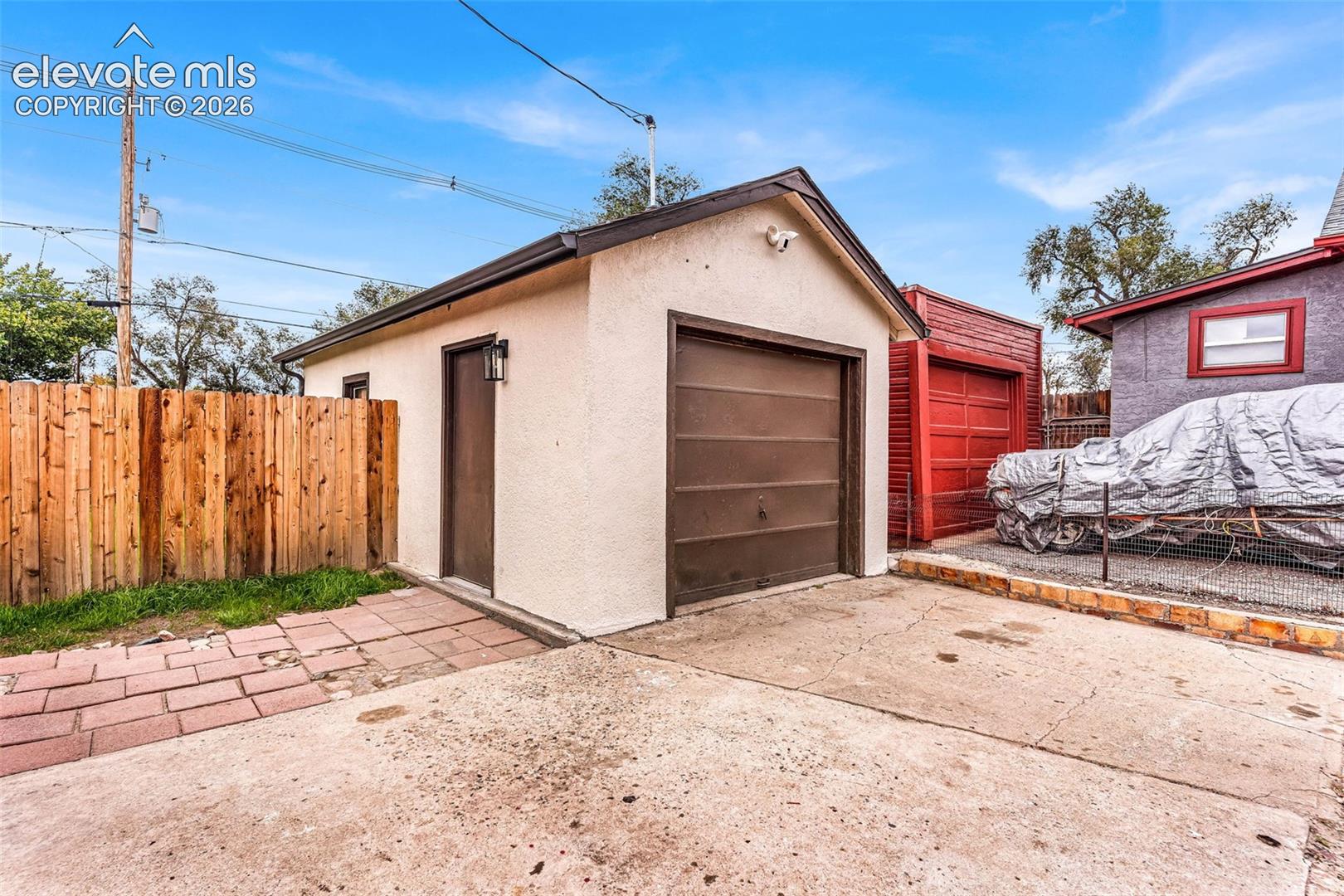 Detached garage featuring stucco siding, a metal overhead door, and a separate side entry door, complemented by a concrete apron and brick paver accents