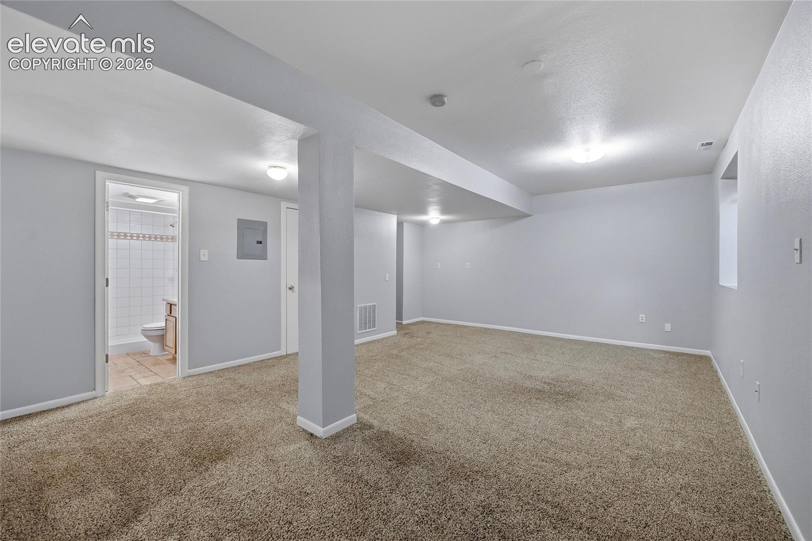 Open-concept room featuring neutral wall-to-wall carpeting, overhead lighting, and a neutral color palette, including a door opening to a tiled bathroom