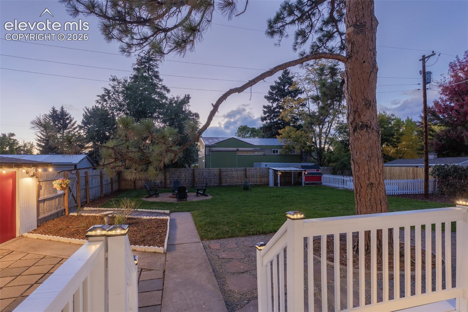 Expansive backyard featuring a manicured lawn, dedicated fire pit area, paved patio, white picket fencing, and mature trees