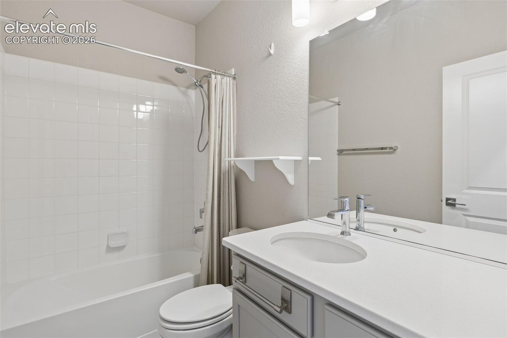 Bathroom featuring a vanity with a white countertop and oval under-mount sink, white tile surround shower-tub combination, large wall-mounted mirror, and wall-mounted shelving