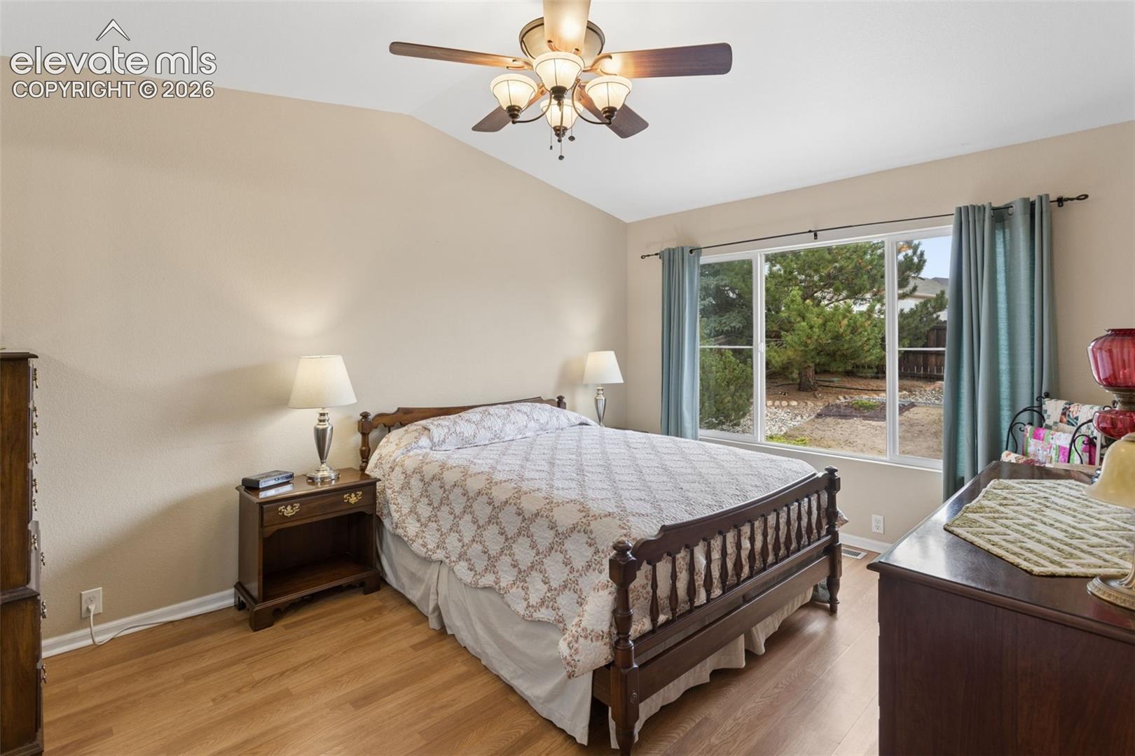 Vaulted ceiling with a ceiling fan light fixture, wood-finish flooring, and a large window with outdoor scenery.