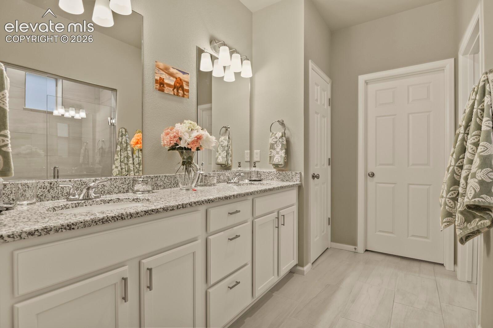 Dual-sink vanity with granite-patterned countertops, white cabinetry, polished metal fixtures, large wall-mounted mirrors, and light-toned tile flooring