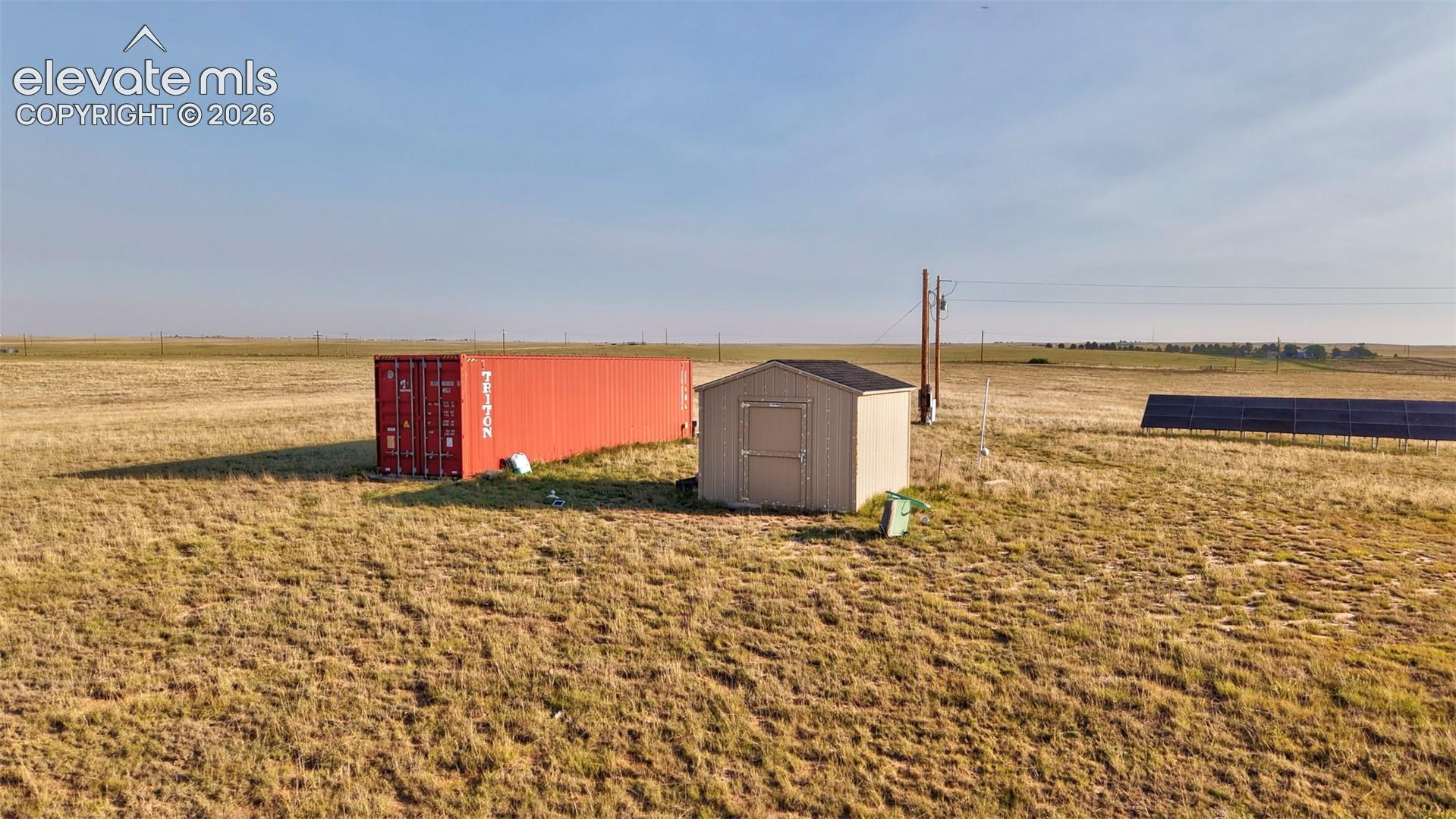 Expansive property featuring a red shipping container, a compact storage shed with a dark roof, and a ground-mounted solar panel array