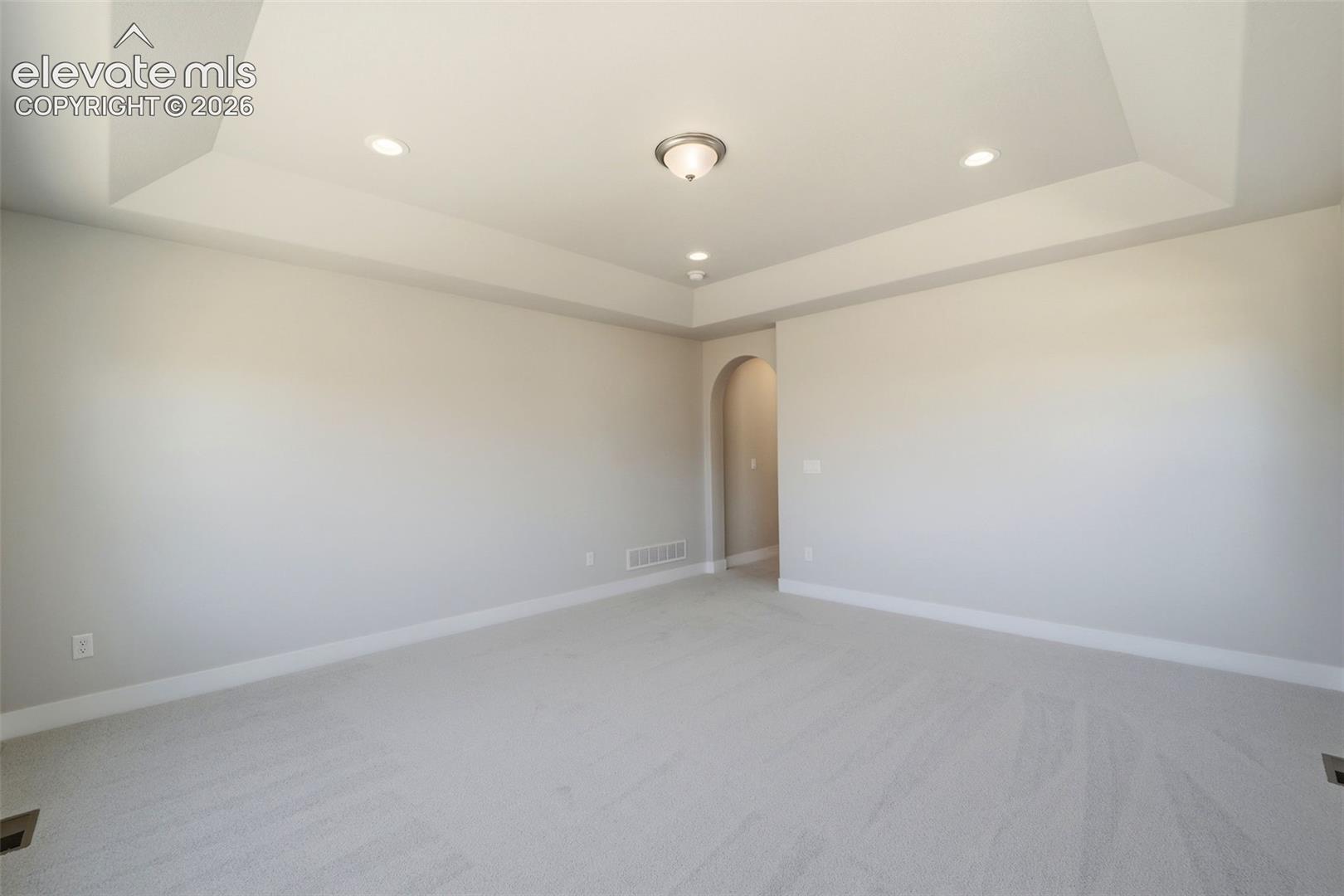 Tray ceiling featuring recessed lighting and a central flush-mount fixture, complemented by neutral wall paint, wall-to-wall carpeting, and an arched entryway