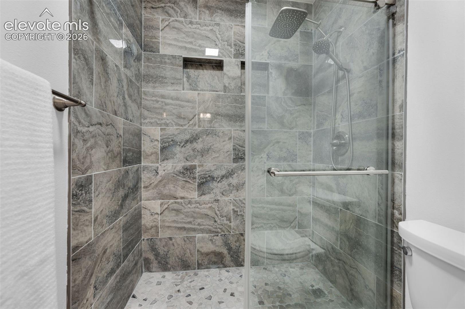 Walk-in shower featuring glass enclosure, large format tile walls, pebble-finish flooring, a built-in bench, and a dual shower head system