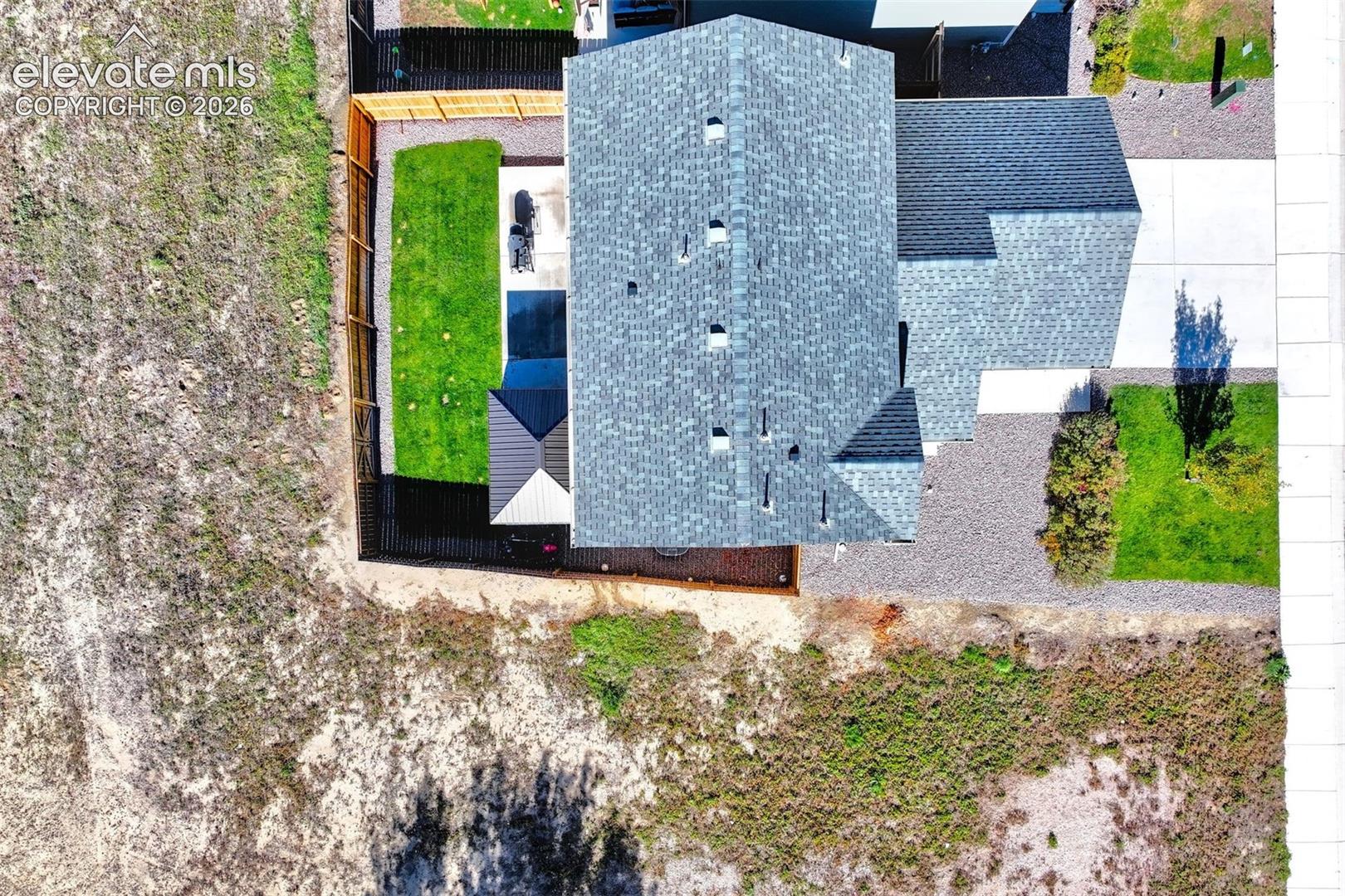 Aerial perspective featuring a multi-pitched asphalt shingle roof, a fenced rear yard with lawn area, a paved driveway, and decorative rock landscaping