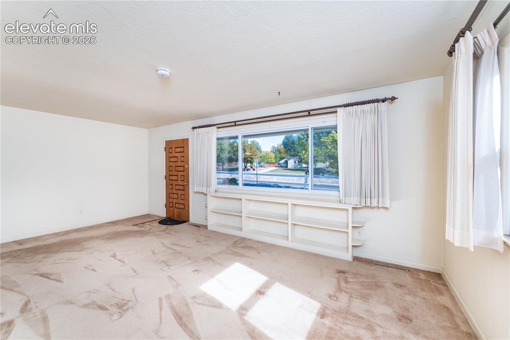 Large window with built-in shelving, patterned wood entry door, and light-colored carpeting