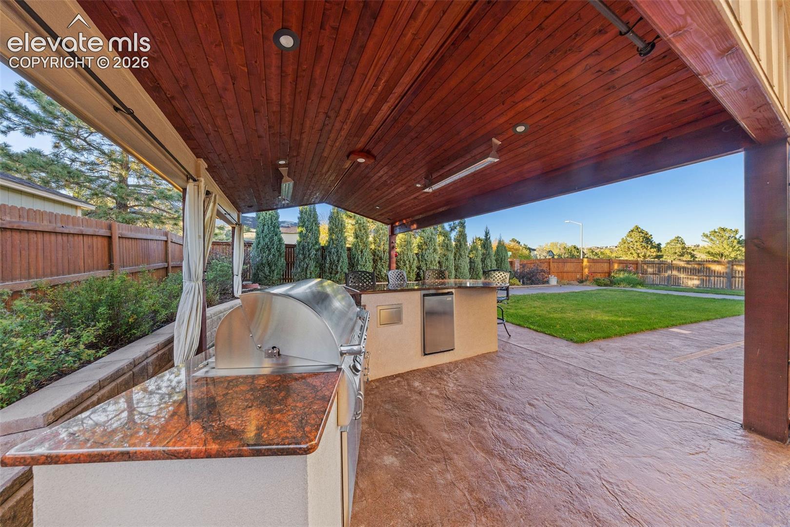 Covered patio featuring a stained wood plank ceiling, stone-textured concrete flooring, and a built-in outdoor kitchen with stone countertop, stainless steel grill, and under-counter refrigerator