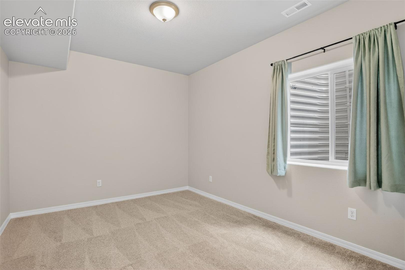 Carpeted room featuring a white-framed window with a horizontal grate, neutral wall paint, white baseboards, and a ceiling light fixture