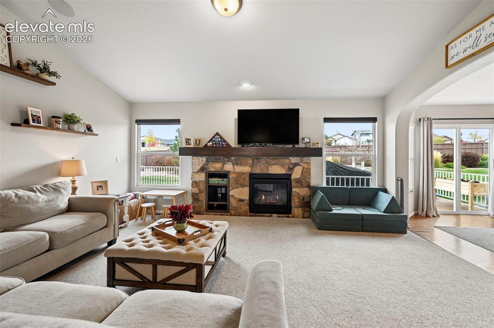 Living area featuring a stone fireplace, a prominent timber mantel, vaulted ceilings, dual-pane windows, and sliding glass doors