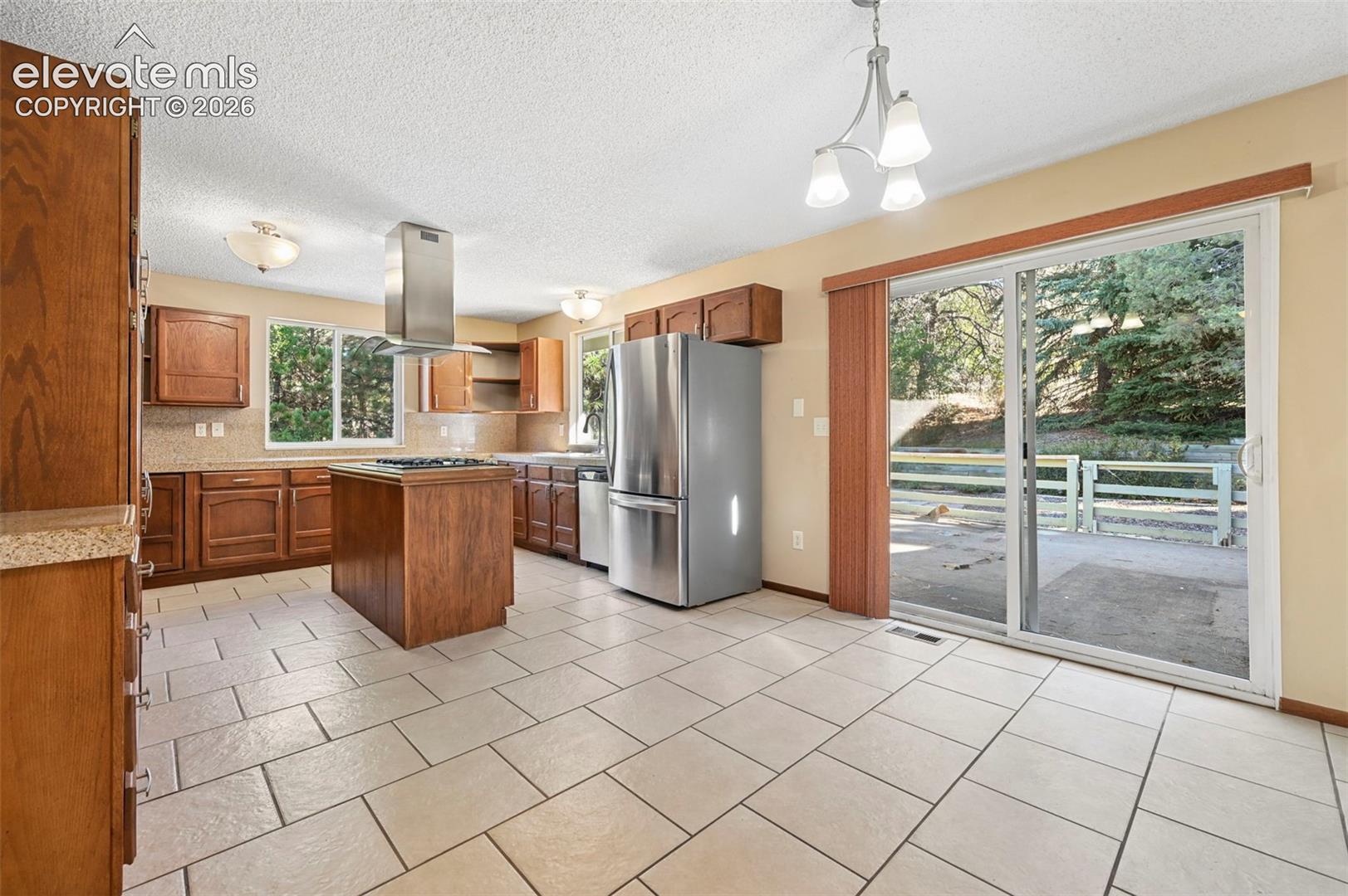 Kitchen featuring wood cabinetry, stainless steel appliances, a central island with an integrated cooktop, a stainless steel range hood, and tile flooring