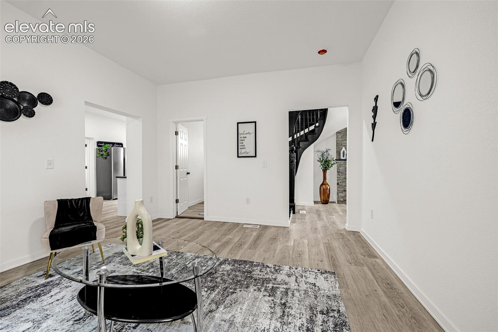 Spacious living area featuring wood-finish flooring, white walls, and a visible entryway to a kitchen area with stainless steel appliances