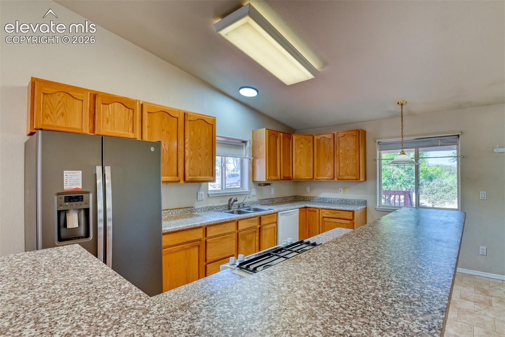 Kitchen featuring natural wood cabinetry, stone-patterned countertops, integrated gas cooktop, double-basin sink, and overhead fluorescent lighting