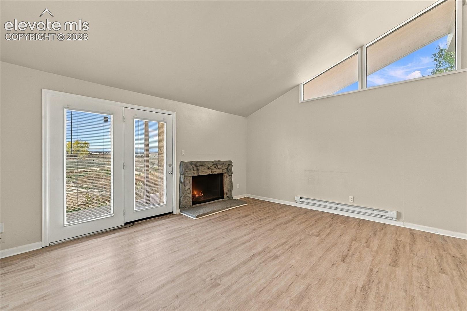 Vaulted ceiling with geometric clerestory windows, wood-finish flooring, stone-surround fireplace, and glass-paneled exterior door