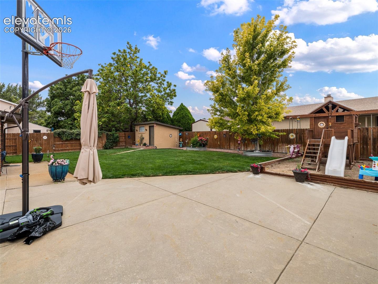 Expansive concrete patio leading to a generous lawn area, featuring a wood storage shed, full perimeter fencing, and mature trees