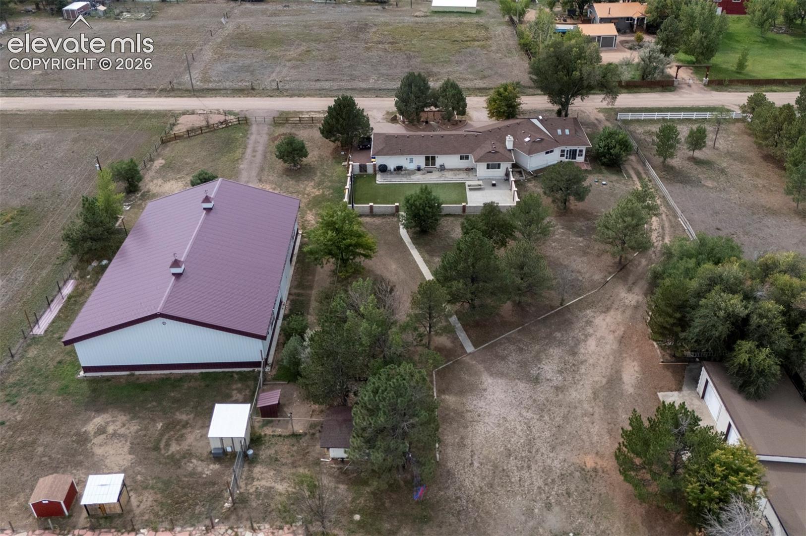 Expansive property featuring a large white residence, separate barn structure with a maroon roof, mature trees, and a paved patio area