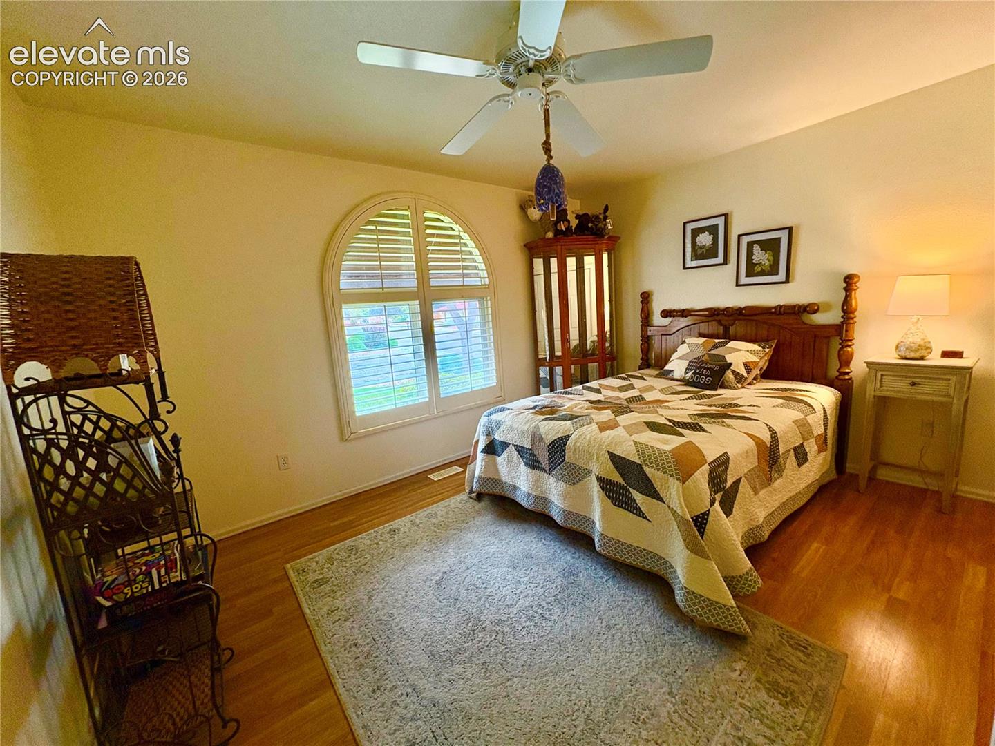 Arch-top window with plantation shutters, ceiling fan with integrated lighting, wood-finish flooring, and sconce lighting