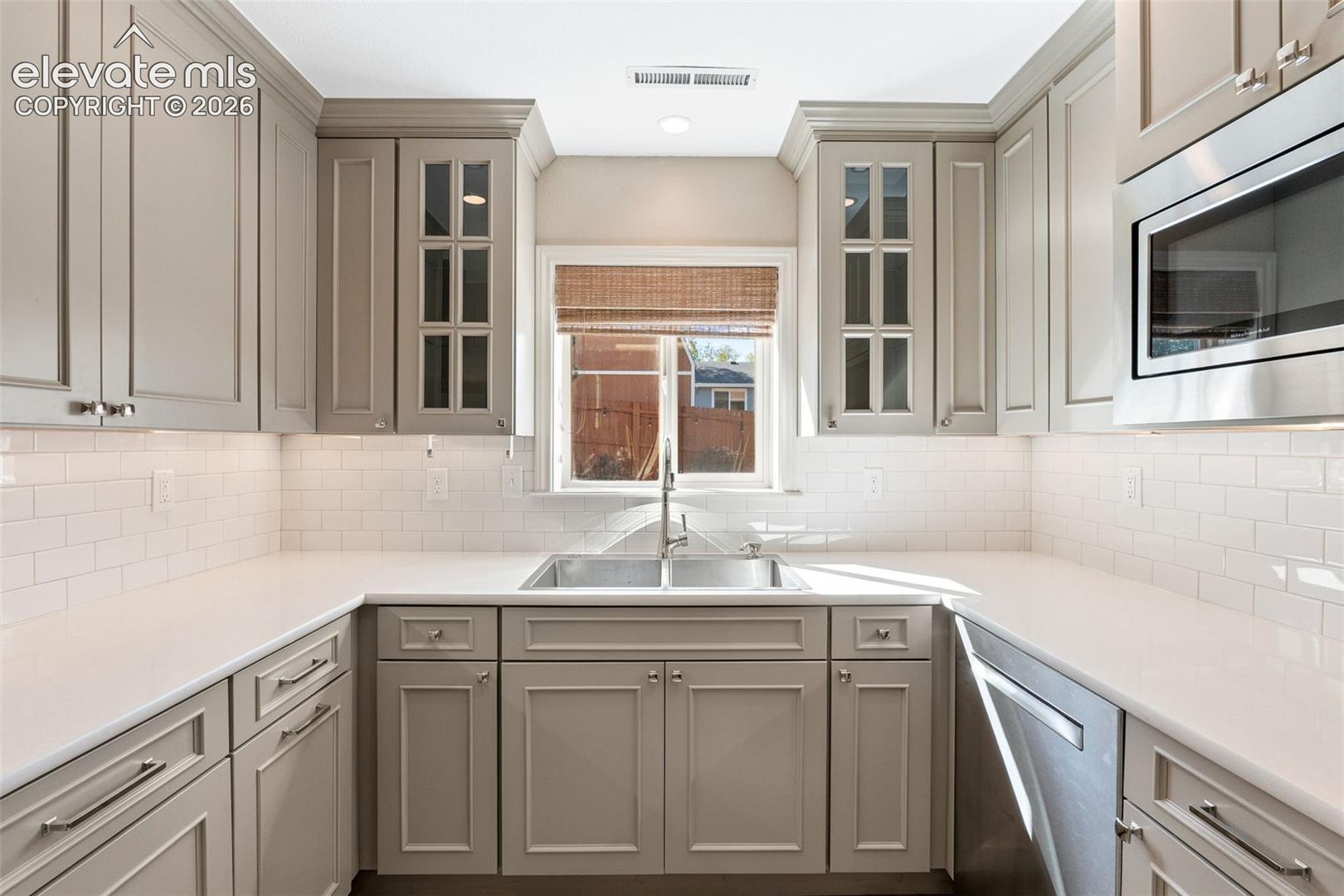 Kitchen featuring shaker-style cabinetry with glass-front accents, solid surface countertops, a white subway tile backsplash, and stainless steel appliances