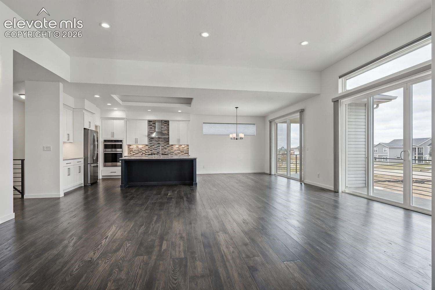 Open-concept living space featuring wide-plank wood-finish flooring throughout, a sleek black kitchen island with pendant lighting, stainless steel appliances, mosaic tile backsplash, white cabinetry, and expansive glass sliding doors opening to a covered balcony with city vistas.  Virtual Stage