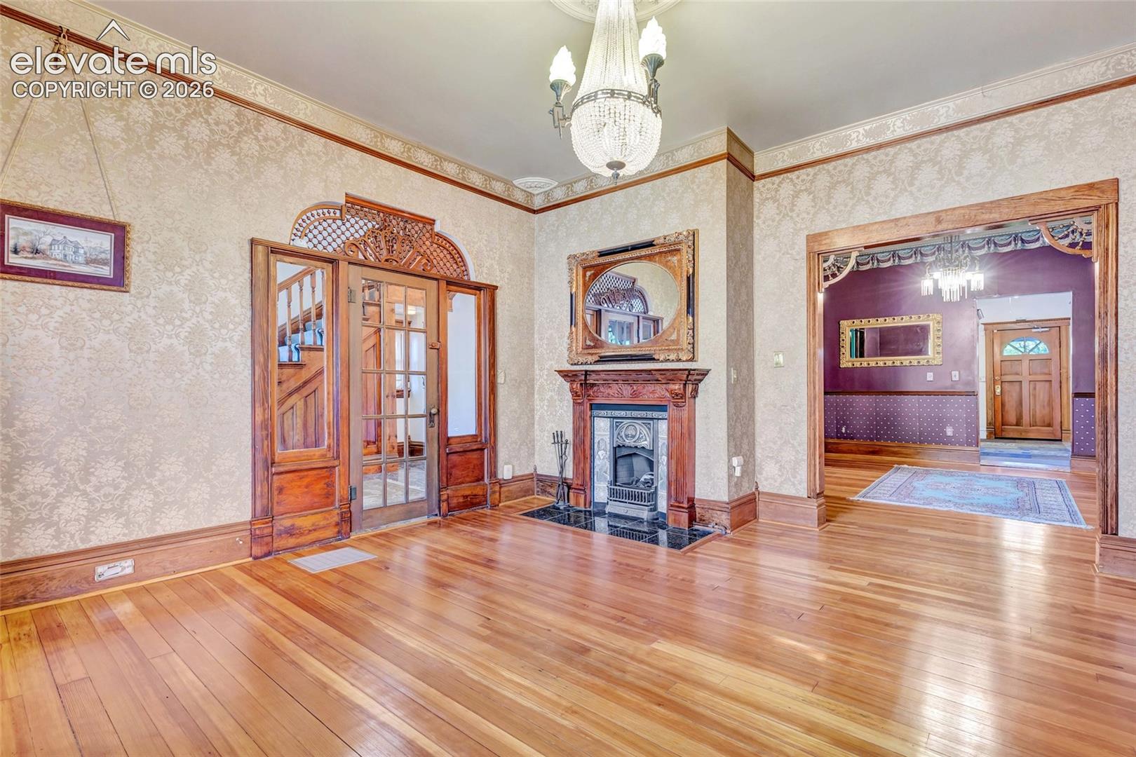 Hardwood flooring, wood-framed glass paneled door with decorative transom, fireplace with wood mantel and tile hearth, ornate ceiling medallion, and crystal-style chandelier