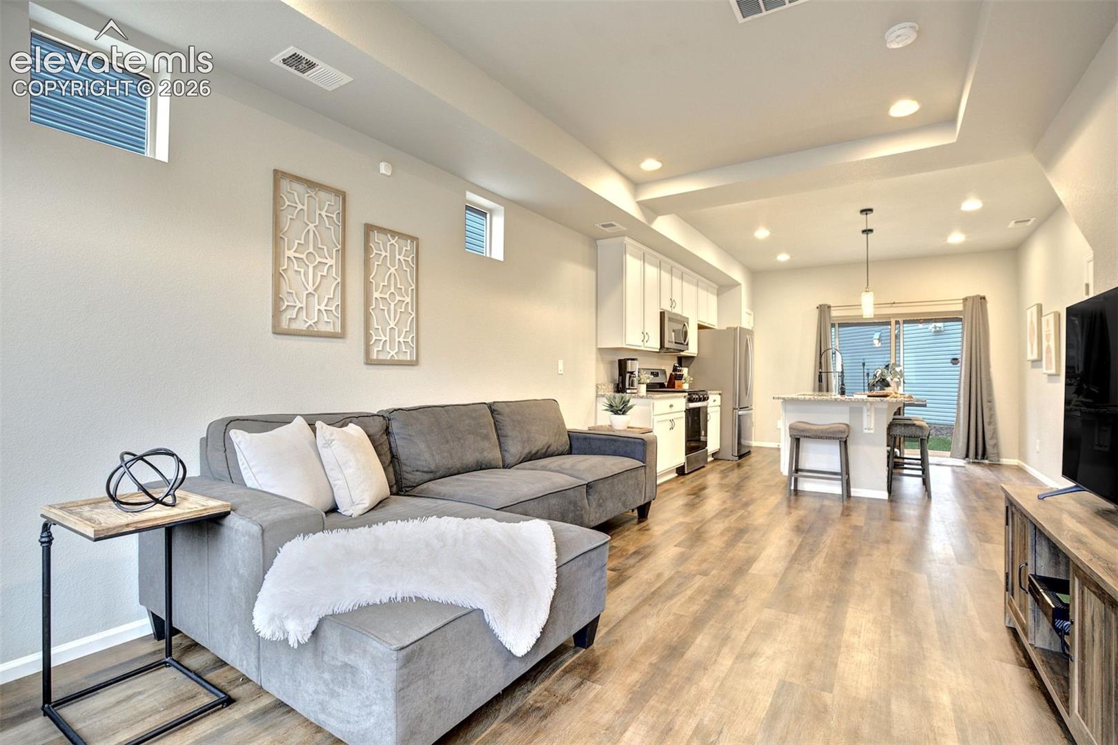 Open-concept interior featuring tray ceilings, recessed lighting, wood-finish flooring, and a kitchen island with stone-finish countertops