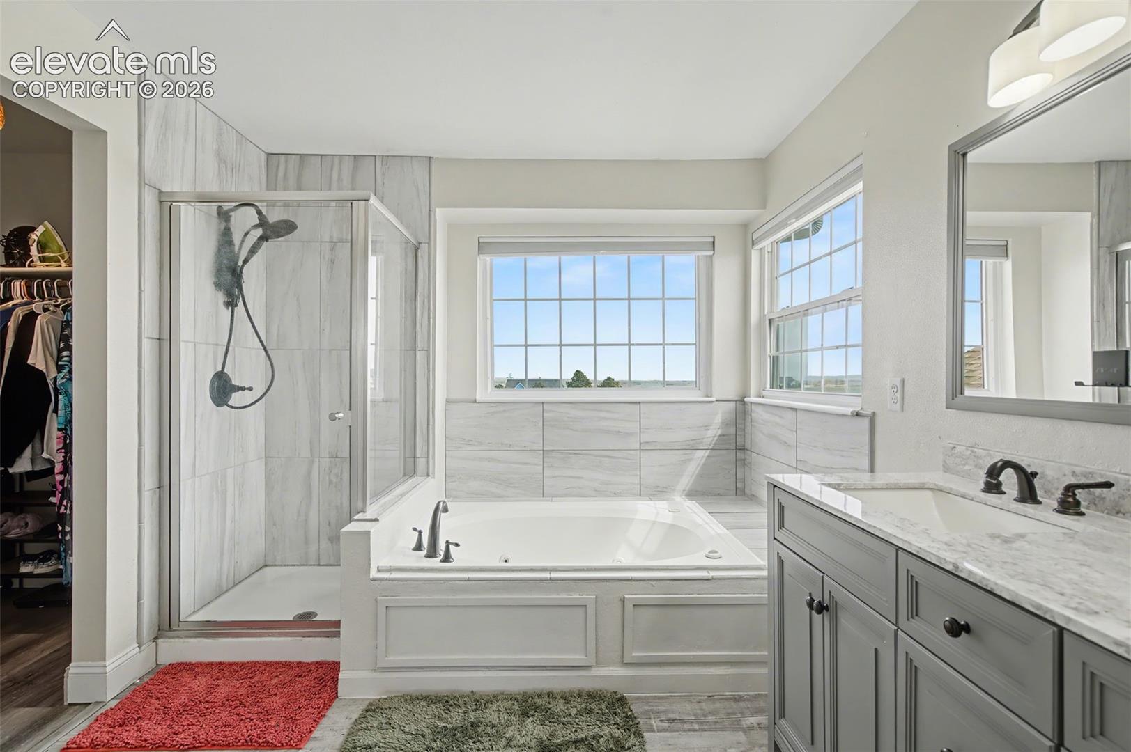 Bathroom featuring a tiled glass-enclosed shower, soaking tub with tile surround, vanity with stone-style countertop, and dual windows