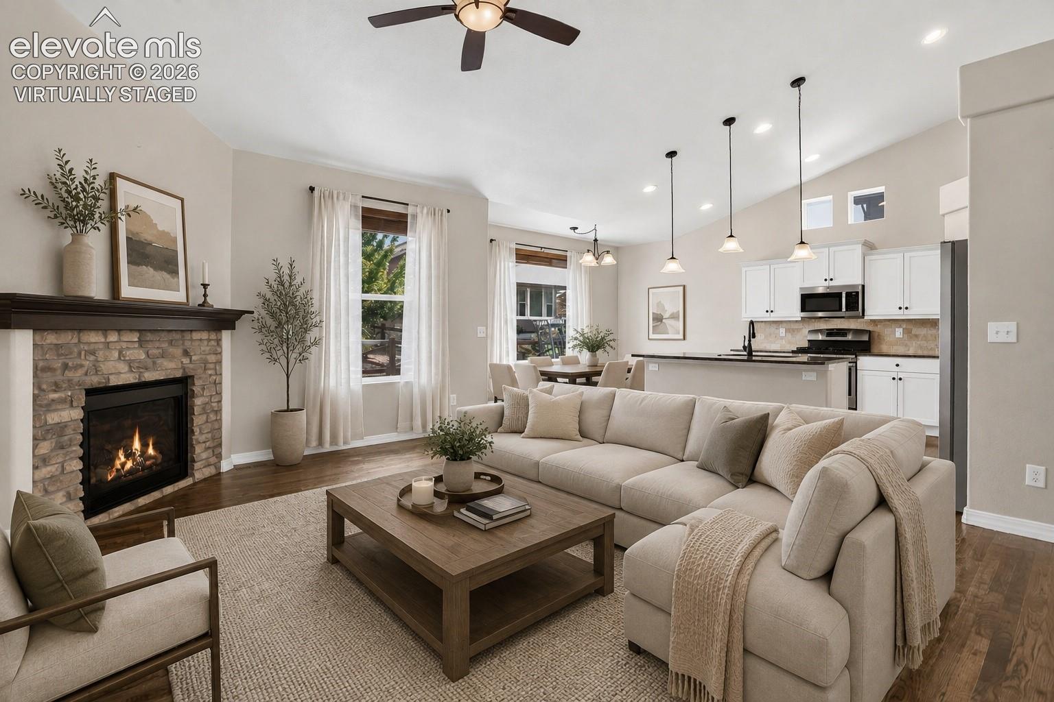 Virtually Staged - Wood-finish flooring, stone-surround fireplace with a dark wood mantel, vaulted ceiling with a ceiling fan, white kitchen cabinetry with pendant lighting, and an integrated kitchen island