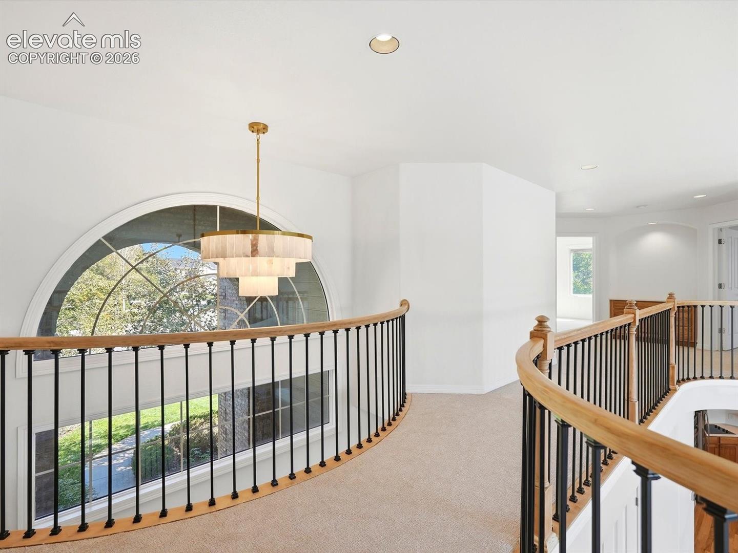 Curved mezzanine gallery featuring a tiered brass chandelier, arched floor-to-ceiling window, and custom railing with wood-finished handrails and metal balusters