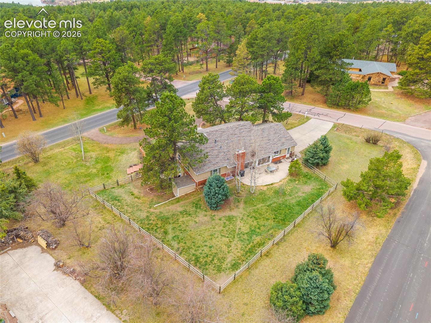 Aerial view featuring a spacious corner lot, masonry chimney, concrete patio, and perimeter fencing, surrounded by mature coniferous trees