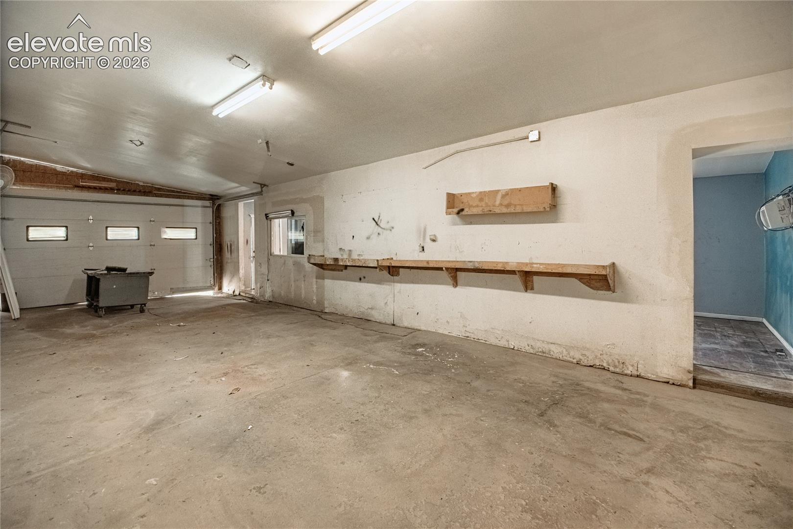 Spacious garage featuring a concrete floor, overhead fluorescent lighting, a roll-up garage door with integrated windows, and built-in wall shelving