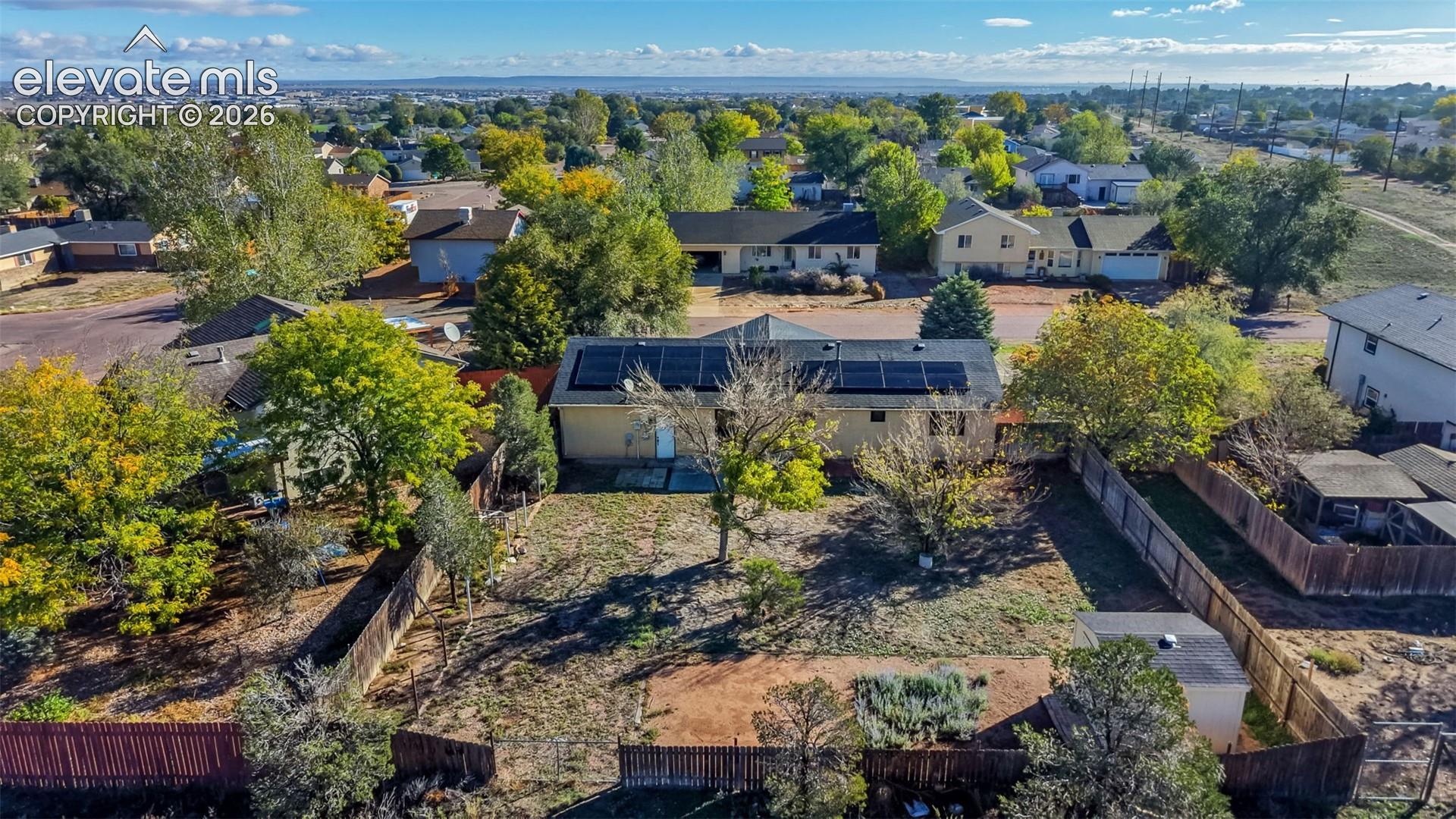 Aerial perspective of property featuring rooftop solar panels, a fenced backyard, mature trees, and a detached storage structure